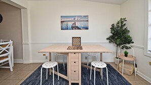 Ein Essbereich mit einem Holztisch, vier weißen Hocker, einem gerahmten Zitat auf dem Tisch, einem Gemälde an der Wand und einer Pflanze in der Ecke.