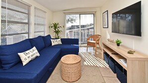 Ein modernes Wohnzimmer mit einem blauen Ecksofa, einem runden Korbstuhl, einem Holz-TV-Möbel und einem großen Fenster mit Jalousien.