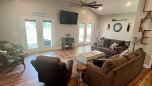 A spacious living room with a ceiling fan, a large sofa, a coffee table, and a television mounted on the wall. - Creekside Cottage-kayaks/fire pit/pet friendly (Jacksons' Gap)
