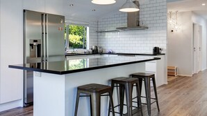 Une cuisine moderne dotée d’un grand îlot central, d’appareils électroménagers en acier inoxydable, de luminaires suspendus et offrant une vue sur la verdure environnante.