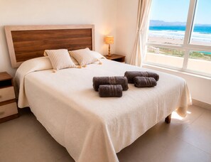 A bedroom with a large bed, a wooden headboard, a nightstand with a lamp, and a view of the beach through the window.