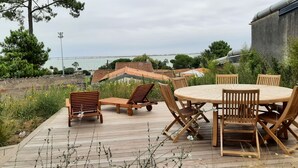 A wooden deck with a round table and chairs, surrounded by greenery and overlooking a residential area with houses and a body of water.