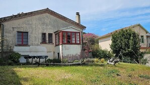 A two-story house with a red window frame, a chimney, and a garden with a table and chairs.