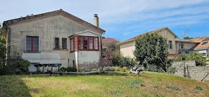 Une maison à deux étages avec un cadre de fenêtre rouge, une cheminée et un jardin agrémenté d’une table et de chaises.