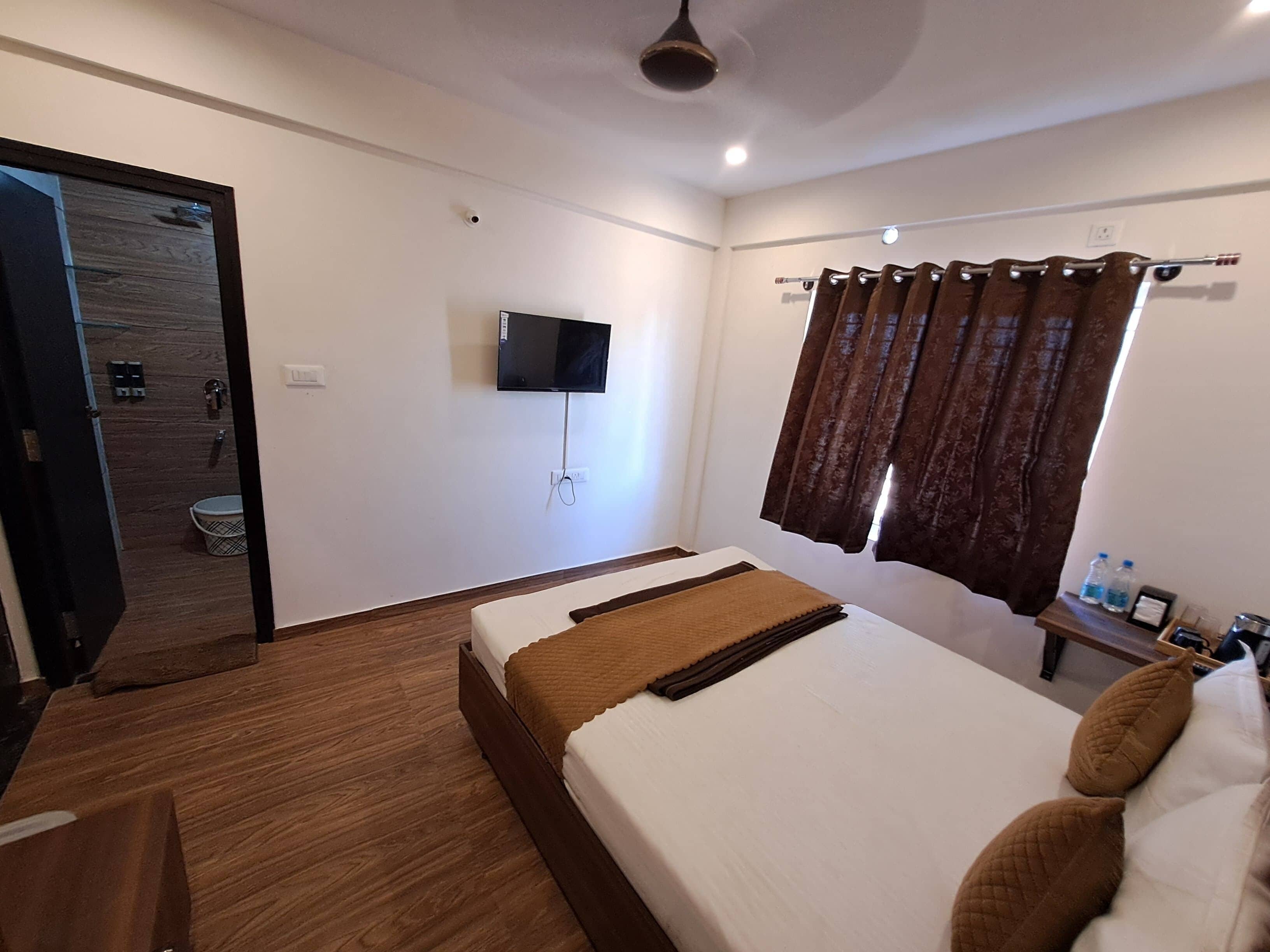 Quarto de hotel com cama, televisão, janela com cortinas, piso de madeira e banheiro visível através de uma porta aberta.