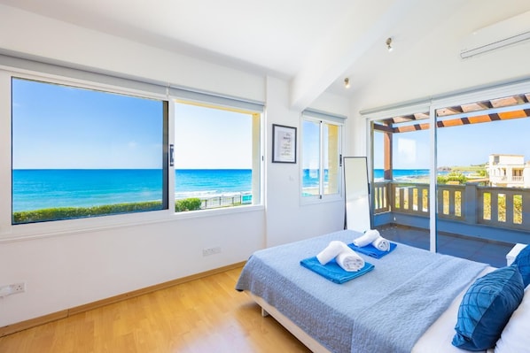 A bedroom with a large window overlooking the sea, a balcony with a view, and a neatly made bed.