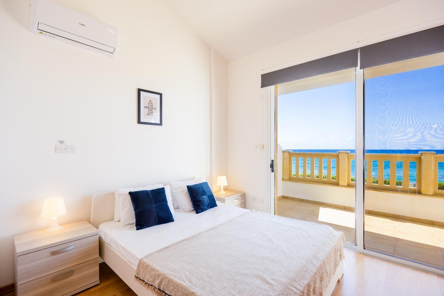 A bedroom with a bed, bedside tables, a framed quote on the wall, and a balcony with a view of the sea.