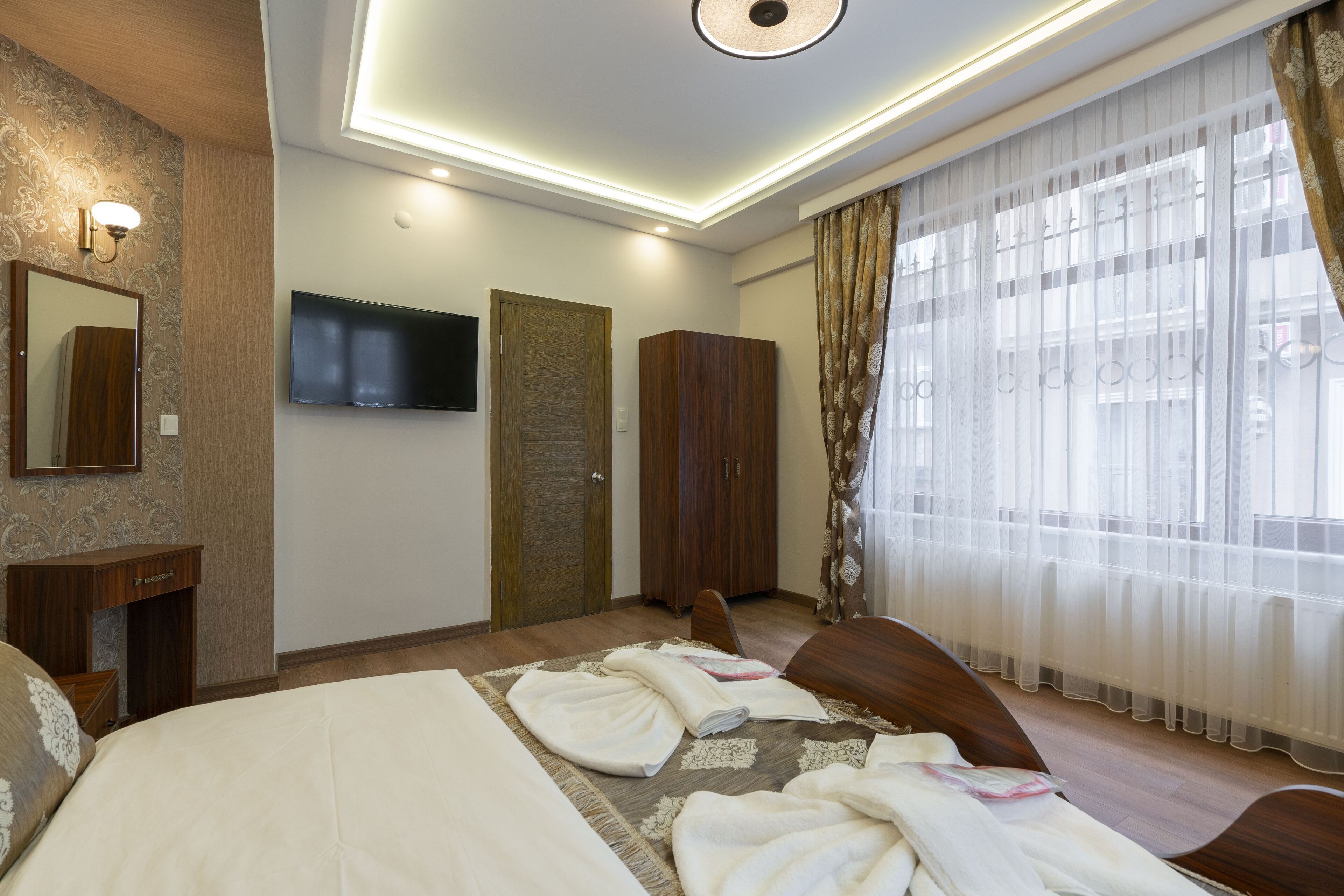 A hotel room with a bed, a wardrobe, a television mounted on the wall, and a window with curtains.