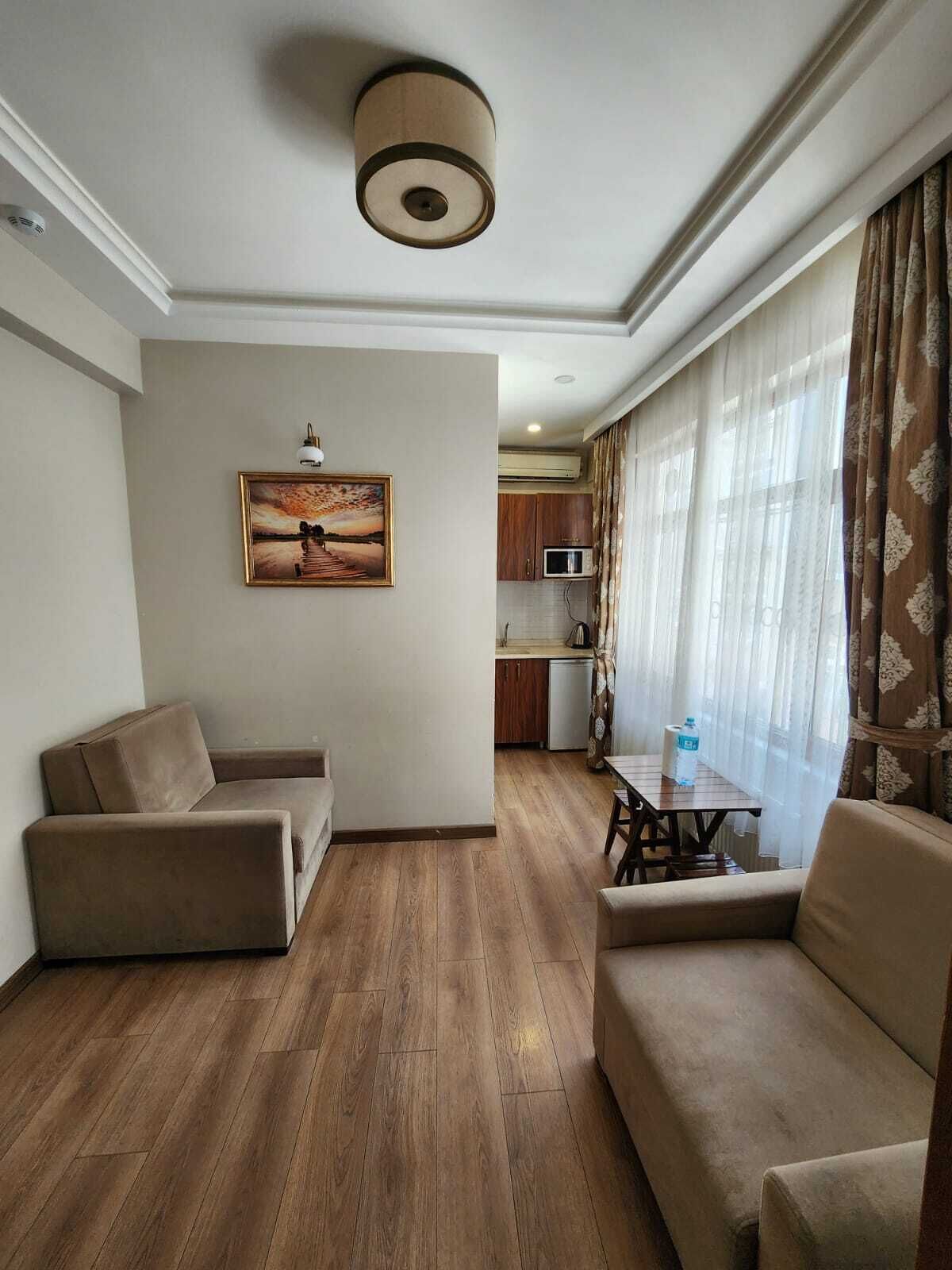 A living room with a sofa, a coffee table, and a painting on the wall. There is a kitchen in the background with a microwave and a water dispenser.