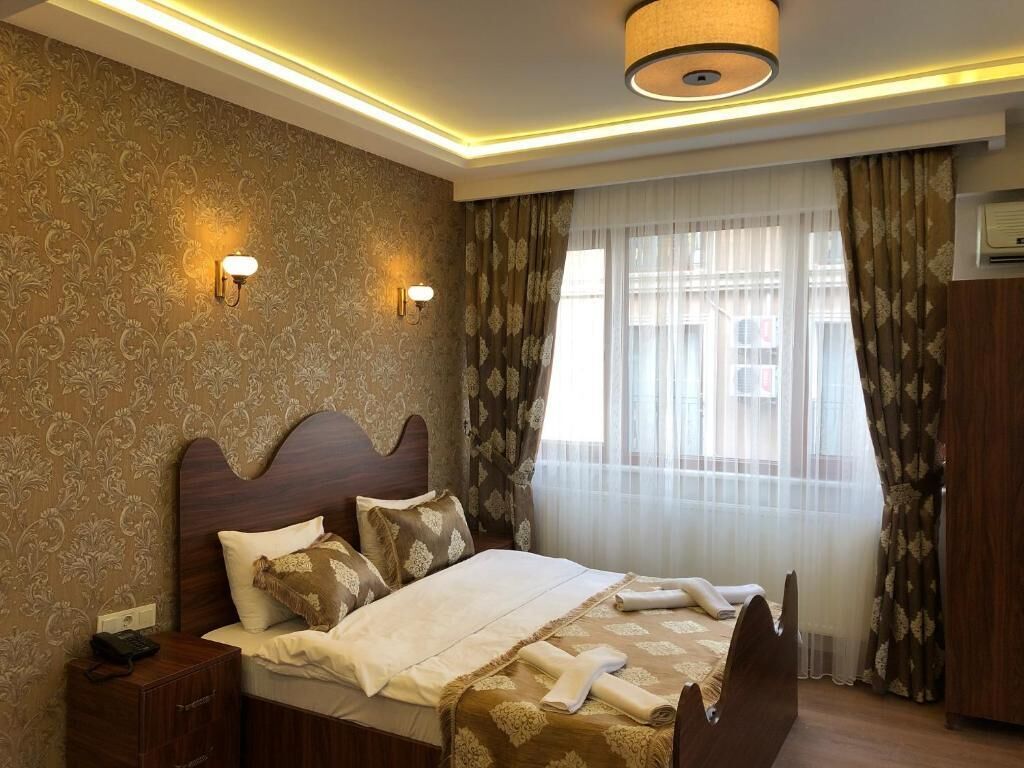 A hotel room with a large bed, two wall-mounted lights, a ceiling light, a window with sheer curtains, and an air conditioning unit.