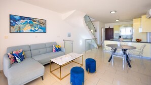 Una sala de estar moderna con un sofá, una mesa de centro y un otomano azul. Una escalera conduce a una cocina con zona de comedor.