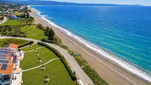 An aerial view of a coastal area with a beach, a road curving along the coast, and a building with solar panels.