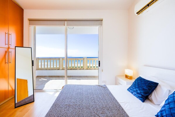 A bedroom with a bed, a nightstand, a lamp, a mirror, and a sliding door leading to a balcony with a view of the sea.