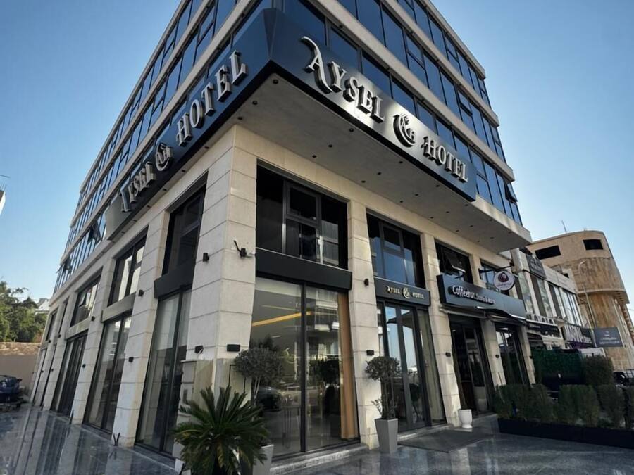 A modern hotel with a prominent 'Aysel & Hotel' sign, large glass windows, and a reflective marble floor.