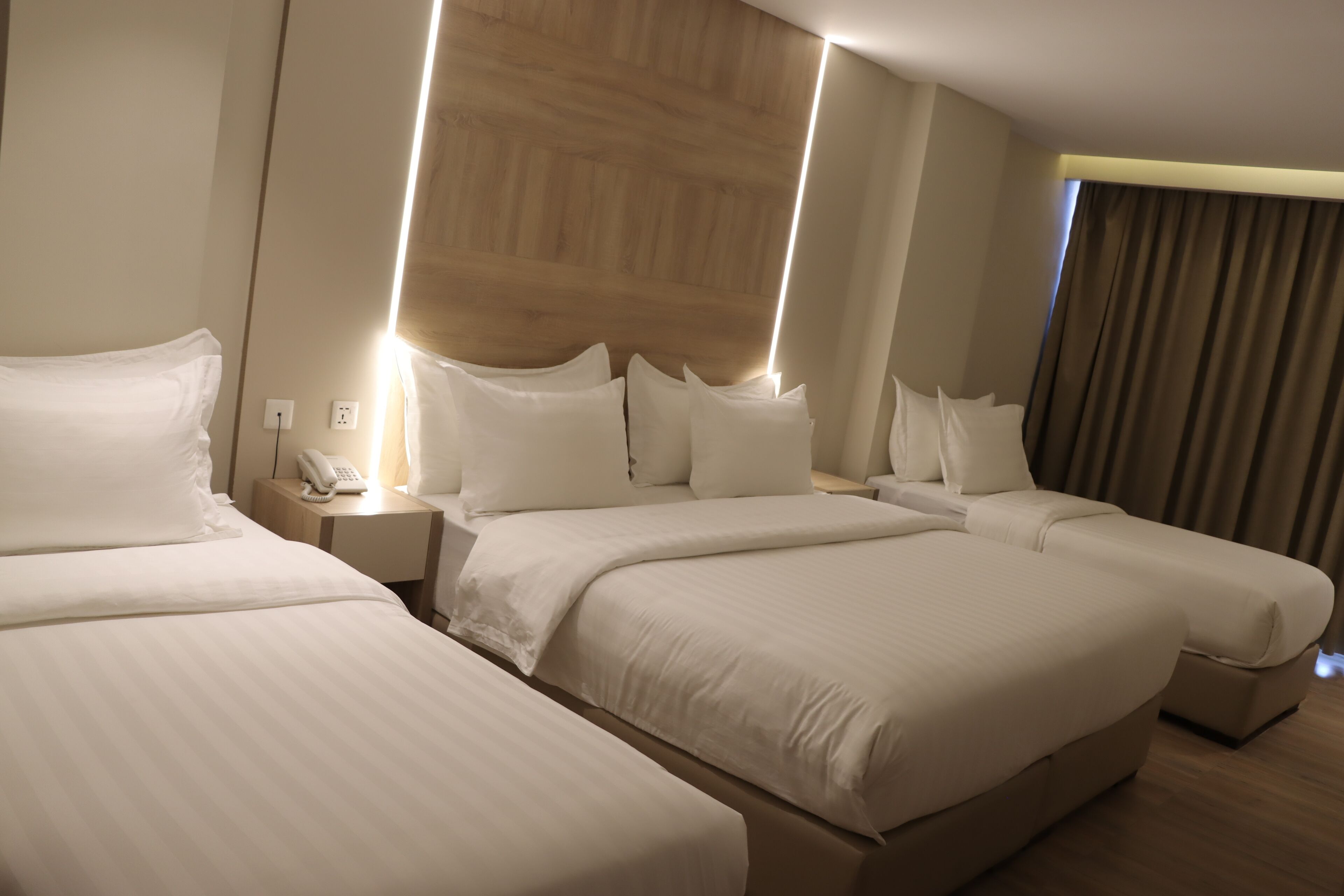 A hotel room with three beds, a wooden headboard, a bedside telephone, and a window with curtains.
