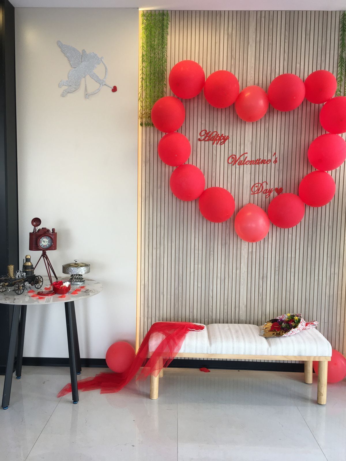 Een kamer met een wand versierd met rode ballonnen in de vorm van een hart, een bankje en een kleine tafel met een klok en decoratieve objecten.