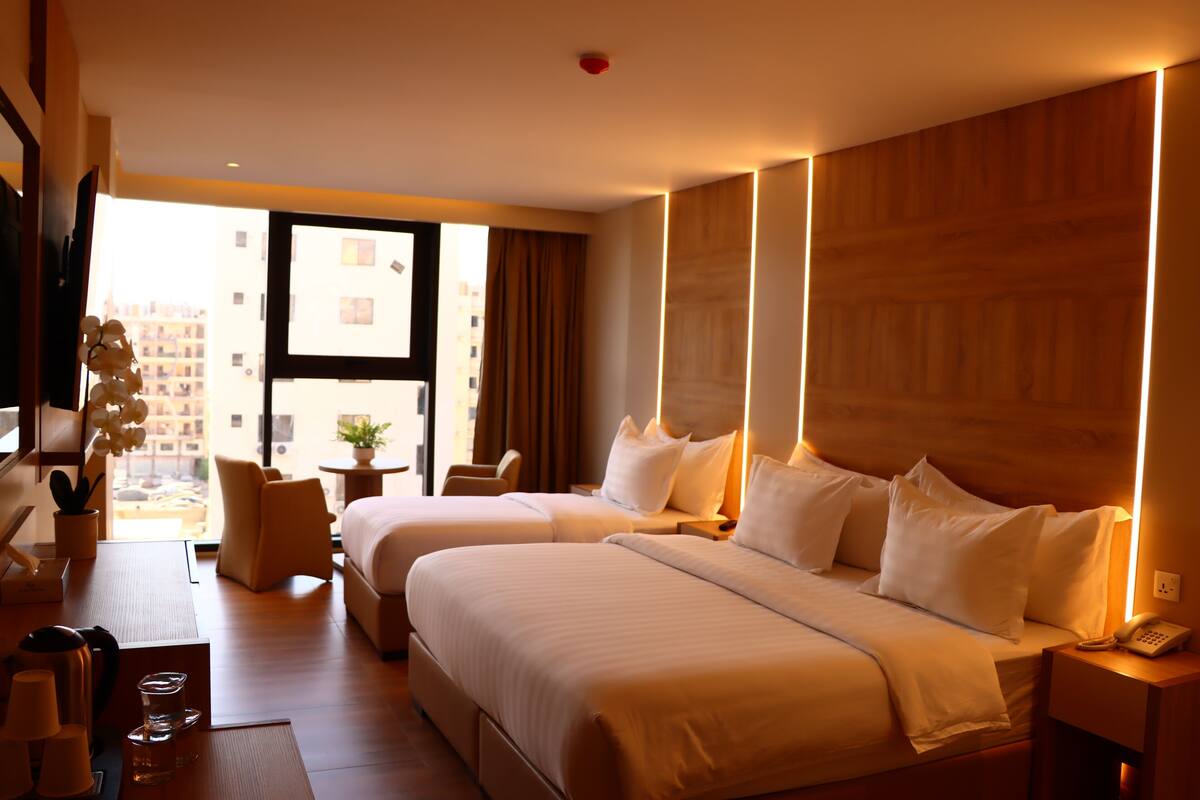 A hotel room with two beds, a desk, a chair, a TV, and a window with a view of buildings.