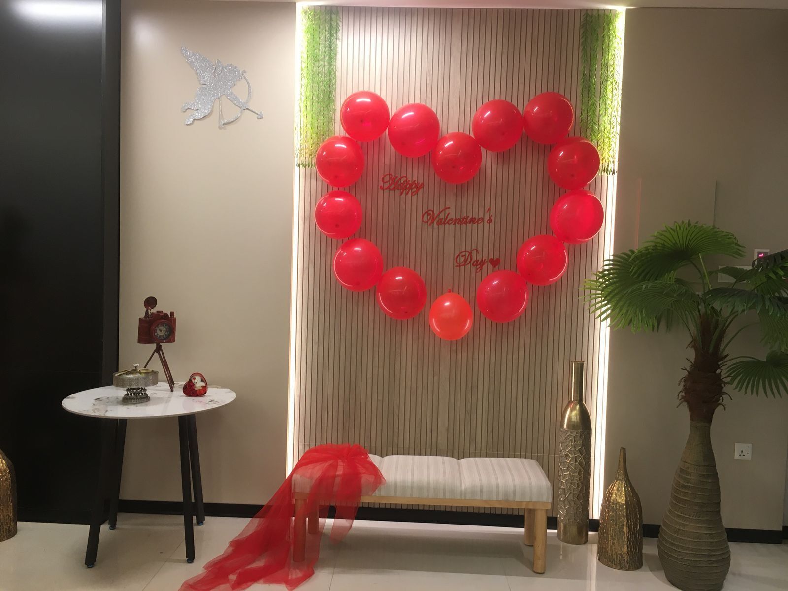 A room with a heart-shaped balloon decoration, a bench, a table with a camera, and a plant.