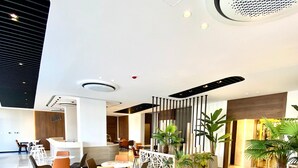 Una moderna hall d'albergo con reception, area salotto con sedie e tavoli, piante decorative e un elemento decorativo a soffitto.