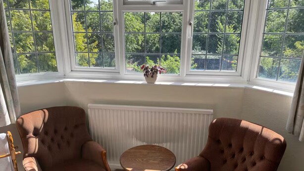 A room with two brown upholstered chairs and a small round table, a white radiator, and a window with a view of greenery. - Private 1-bedroom apartment in Raynes Park (London)