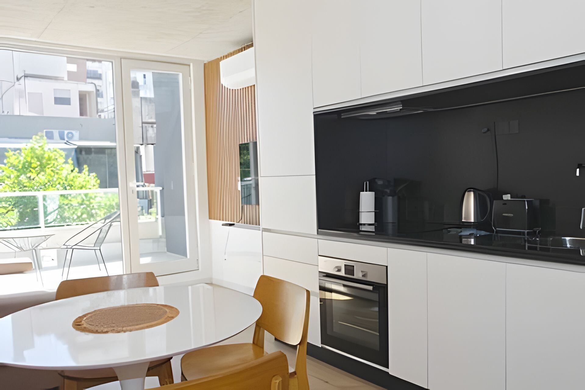 Cocina moderna con armarios blancos, encimera negra y electrodomésticos de acero inoxidable. Cuenta con una zona de comedor con una mesa blanca y sillas de madera.