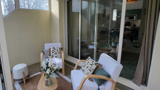 Eine sonnendurchflutete Terrasse mit einer Glasschiebetür zu einer Küche. Zwei weiße, gepolsterte Stühle und ein kleiner Tisch mit einem Blumenstrauß sind zu sehen.