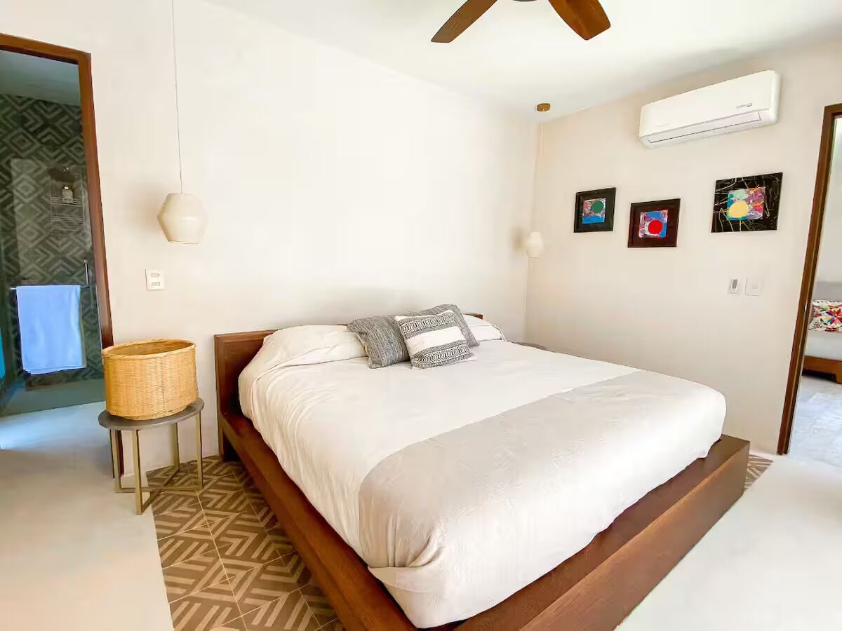 A bedroom with a bed, a wall-mounted air conditioner, and three framed artworks on the wall.