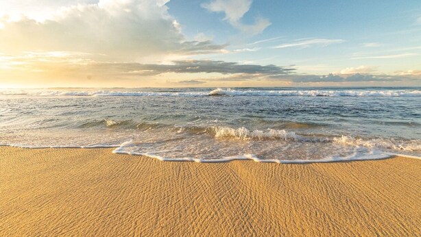 Une plage où les vagues viennent doucement lécher le rivage sous un ciel partiellement nuageux.