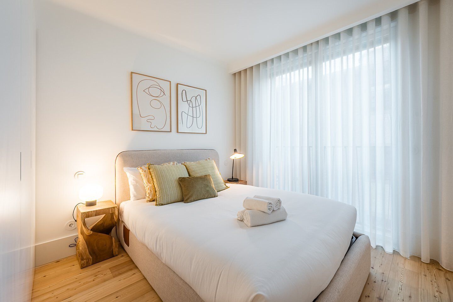 A neatly arranged bedroom with a large bed, two bedside lamps, and a window with sheer curtains.