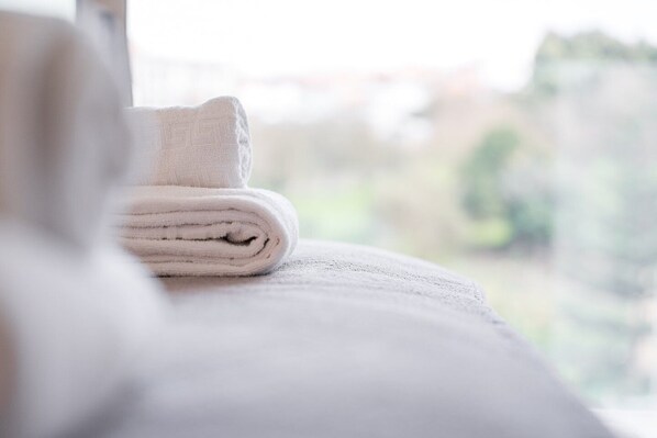 Une serviette blanche, soigneusement pliée, est posée sur une surface blanche, peut-être un lit ou un tapis de bain, et on aperçoit, flou, de la verdure à l’extérieur.