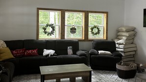 Una sala de estar con ventilador de techo, un sofá, una silla, una mesa de centro y una alfombra. Hay tres ventanas y coronas decorativas en la pared.