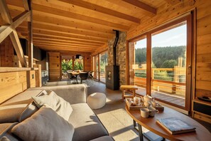 Interior acollidor d'una cabana amb sostre de fusta, una llar de pedra, un sofà, una taula de menjador amb cadires i grans finestres que ofereixen vistes a l'exterior.