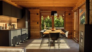 A modern kitchen with a wooden ceiling, a dining area with a large table and chairs, and a view of greenery through the windows.