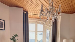 Een kamer met een houten plafond, een kroonluchter, een potplant, een staande lamp en een uitzicht door een groot raam.