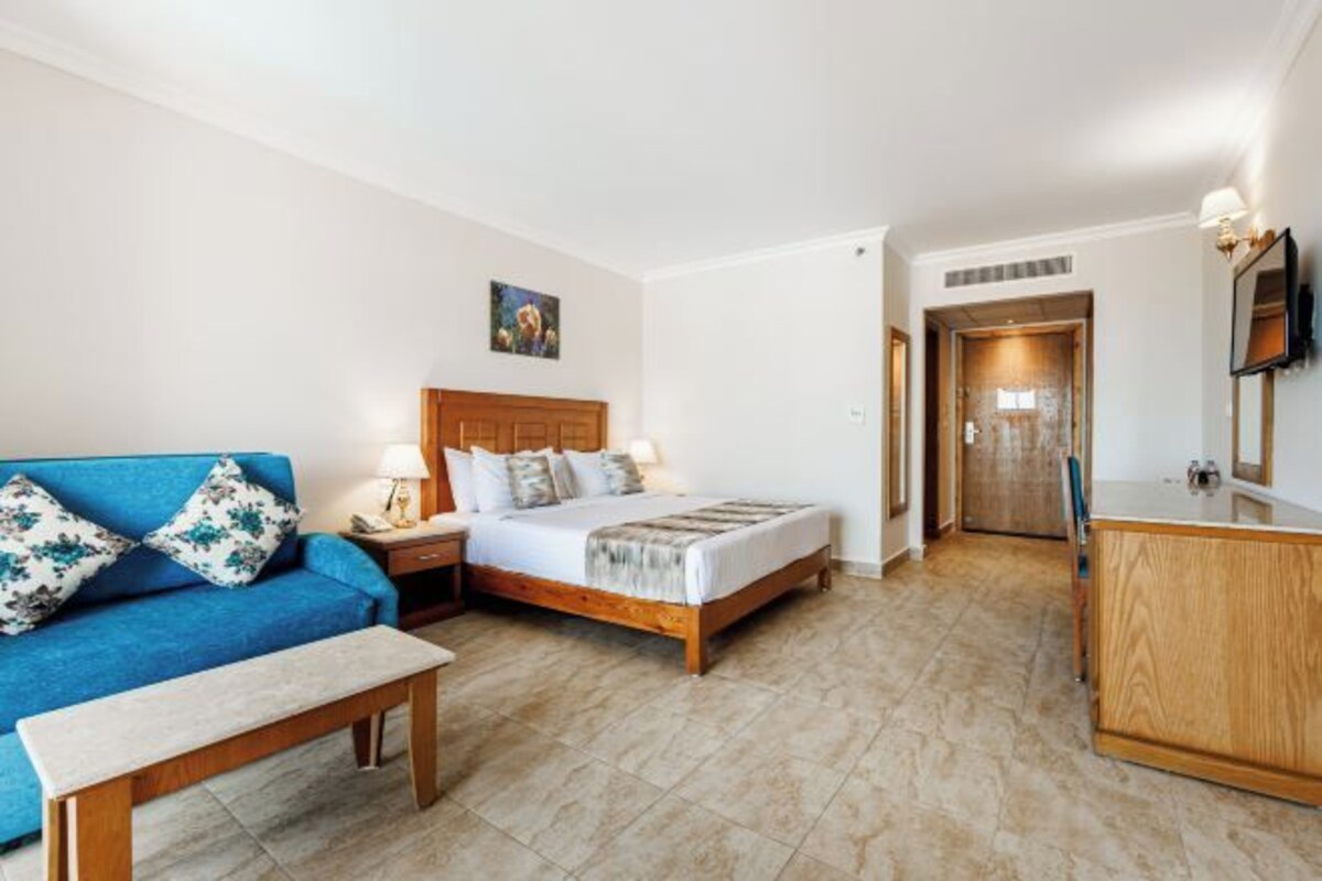 Une chambre d’hôtel avec un lit, un canapé bleu, une table de chevet en bois, une commode en bois et un miroir.