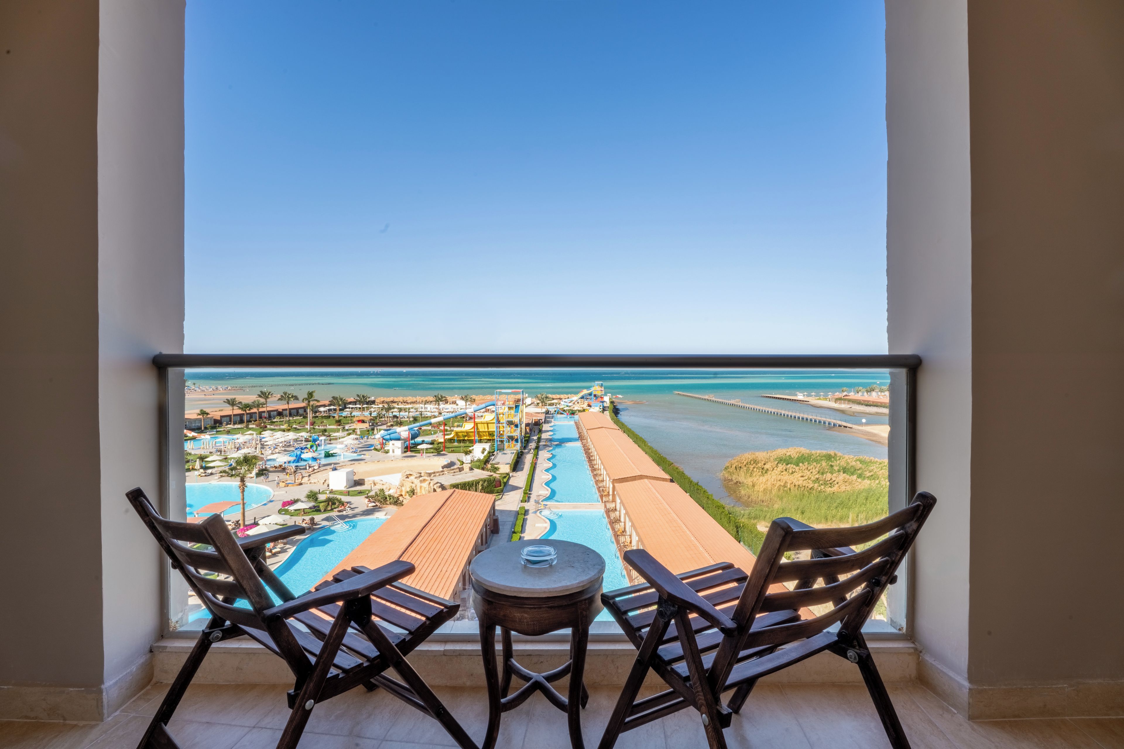 Un balcon avec deux chaises en bois et une petite table, donnant sur un complexe hôtelier doté d’une piscine, de toboggans aquatiques et d’une plage.