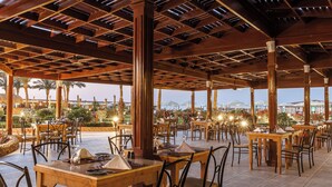 An outdoor dining area with wooden tables and chairs, a tiled floor, and a view of palm trees and buildings in the background. - SUNRISE Solara Aqua park Resort (Hurghada)