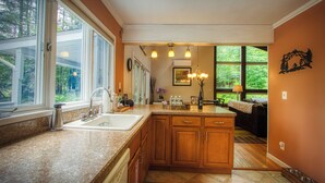 A kitchen with a large window, wooden cabinets, a sink, and a dining area visible through the window. - 3-Bedroom 2 Bath Cozy Chalet 1 Mile From Mountain! (Lanesville)
