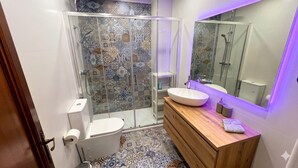 Une salle de bain avec une douche à parois de verre, des toilettes blanches, un meuble-lavabo en bois doté d’un grand évier et un miroir.