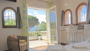 Un dormitorio con cama, una silla de mimbre, un escritorio con sillas y un balcón con vistas a árboles y una masa de agua.