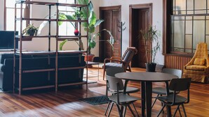 Um quarto espaçoso com piso de madeira, uma mesa redonda com cadeiras, um sofá preto, uma poltrona amarela e uma estante de madeira.