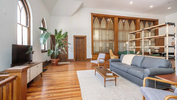 Uma sala de estar ampla com piso de madeira, um sofá cinza, uma mesa de centro de madeira e uma planta grande em vaso.