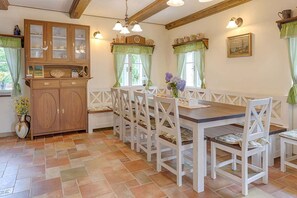 Ein Essbereich mit Holztisch, weißen Stühlen, einem Holzkabinett mit Glastüren und grünen Vorhängen an den Fenstern.
