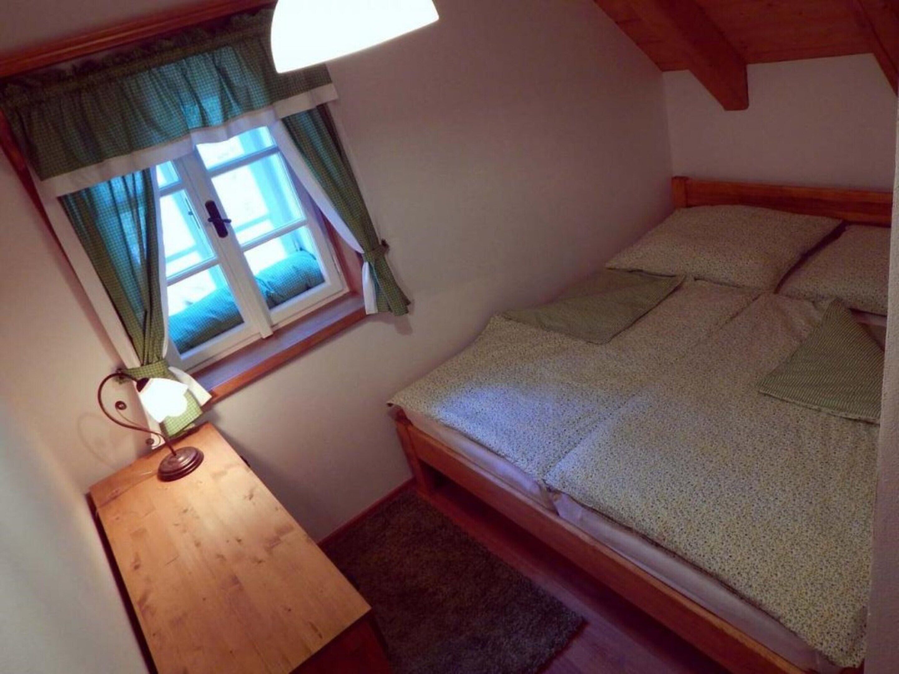 A small, single-bed room with a wooden headboard, a bedside table with a lamp, and a window with curtains.