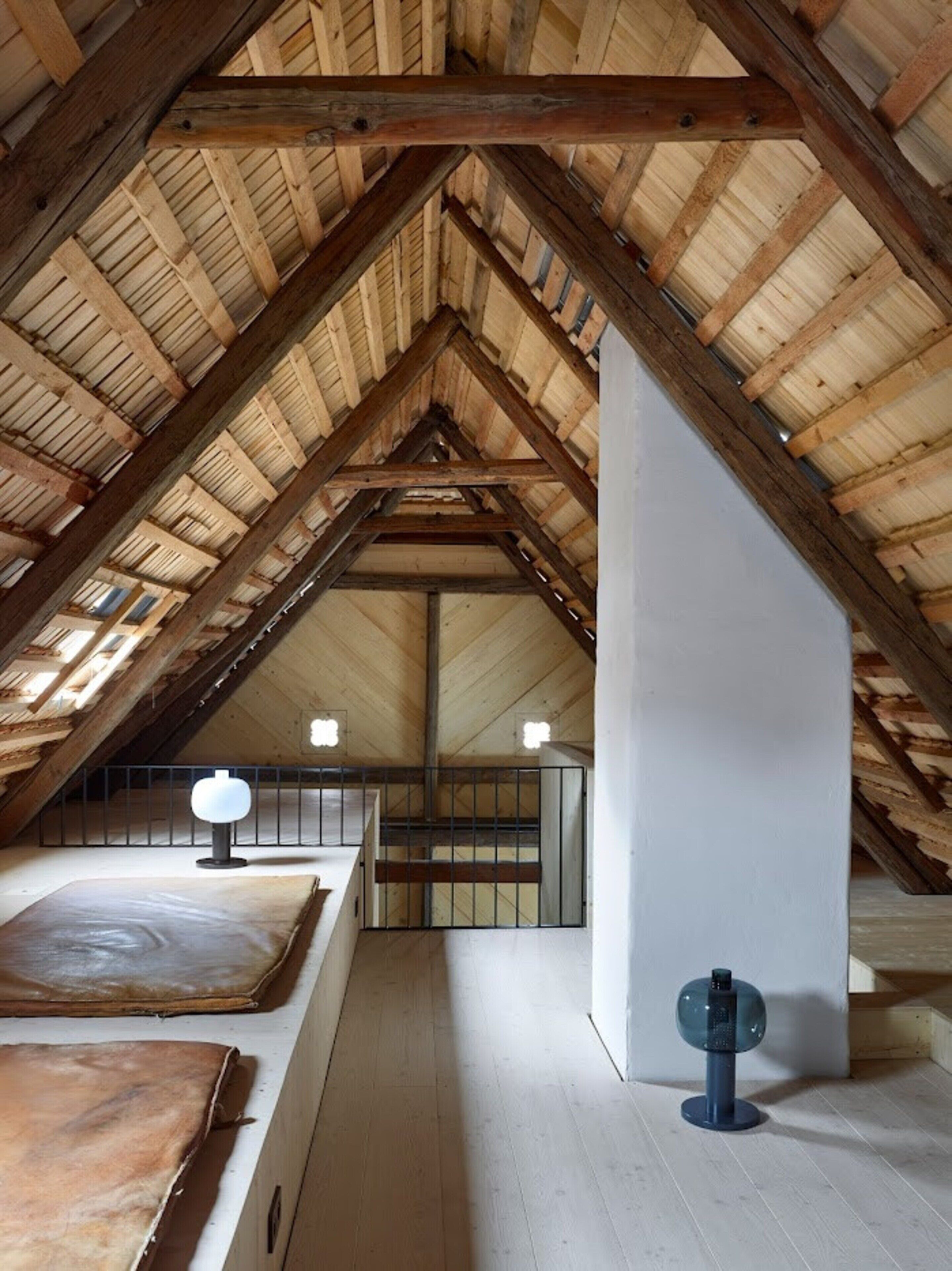 Habitación en ático con techo de madera inclinado, pared blanca y una pequeña mesa con lámpara.