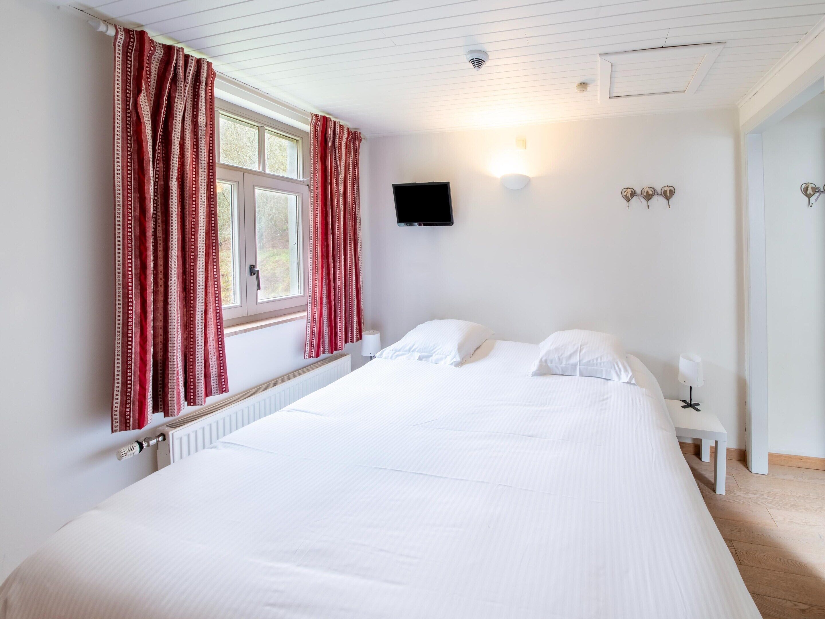 A bedroom with a large bed, a window with red curtains, a wall-mounted TV, and a bedside table with a lamp.