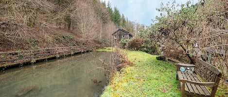 A wooden bench by a calm river, surrounded by dense trees and a rustic building in the background.