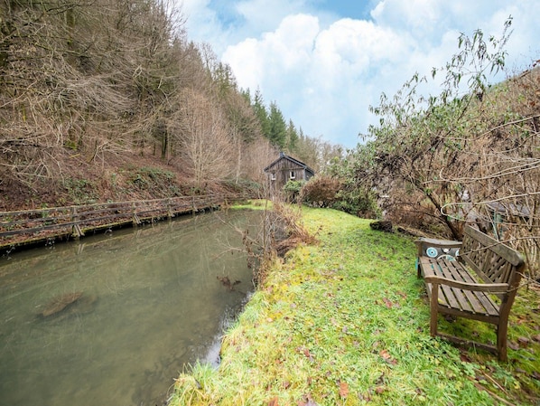 Una panchina di legno vicino a un fiume tranquillo, circondata da alberi rigogliosi e un edificio rustico sullo sfondo.