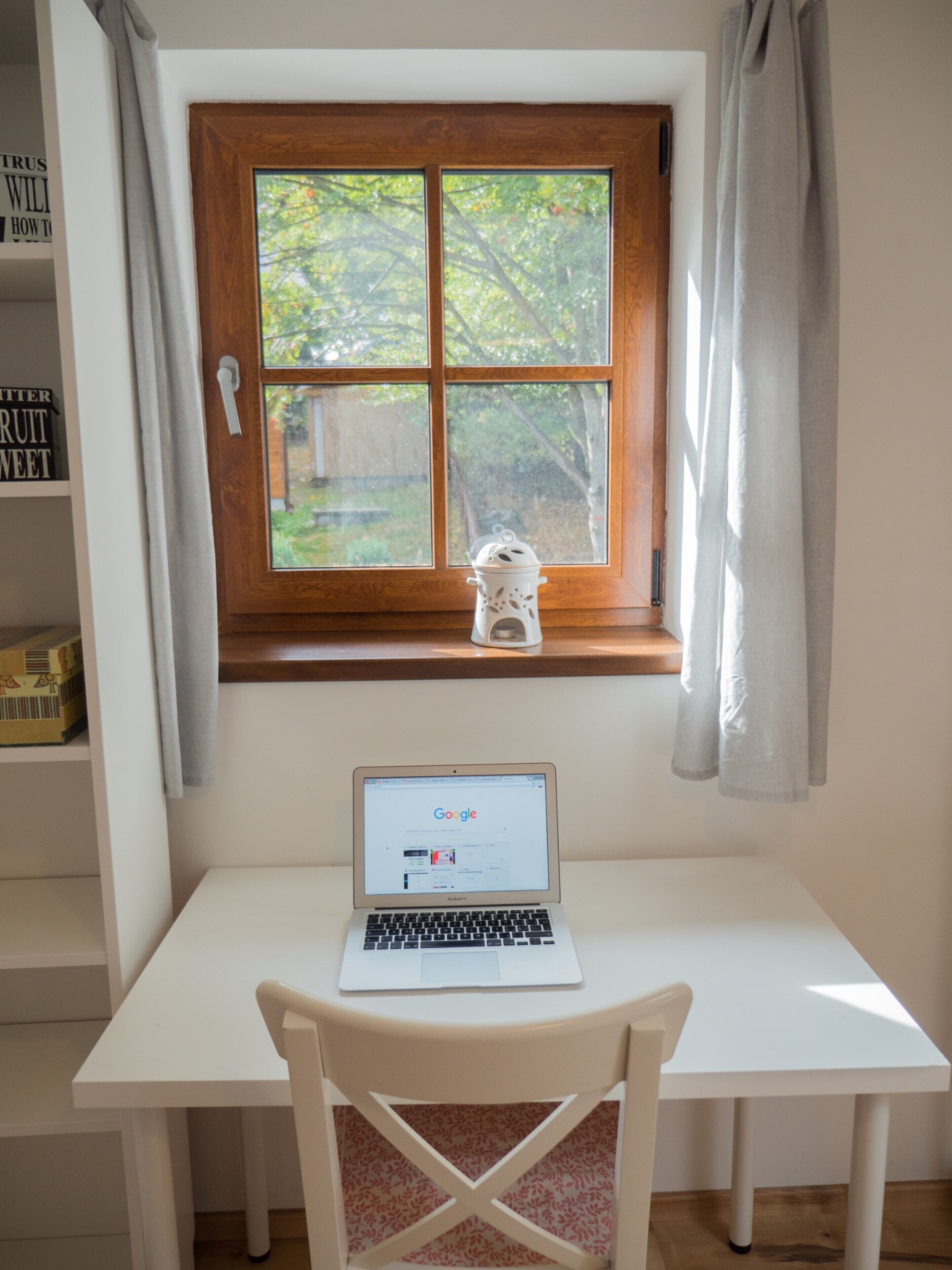 A white desk with a laptop, a window with a wooden frame, and a white chair.