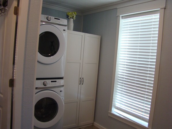 A laundry room with two stacked front-loading washing machines, a white cabinet, and a window with blinds. - Charming 2 bedroom home in Tallassee, Alabama near Lake Martin (Tallassee)